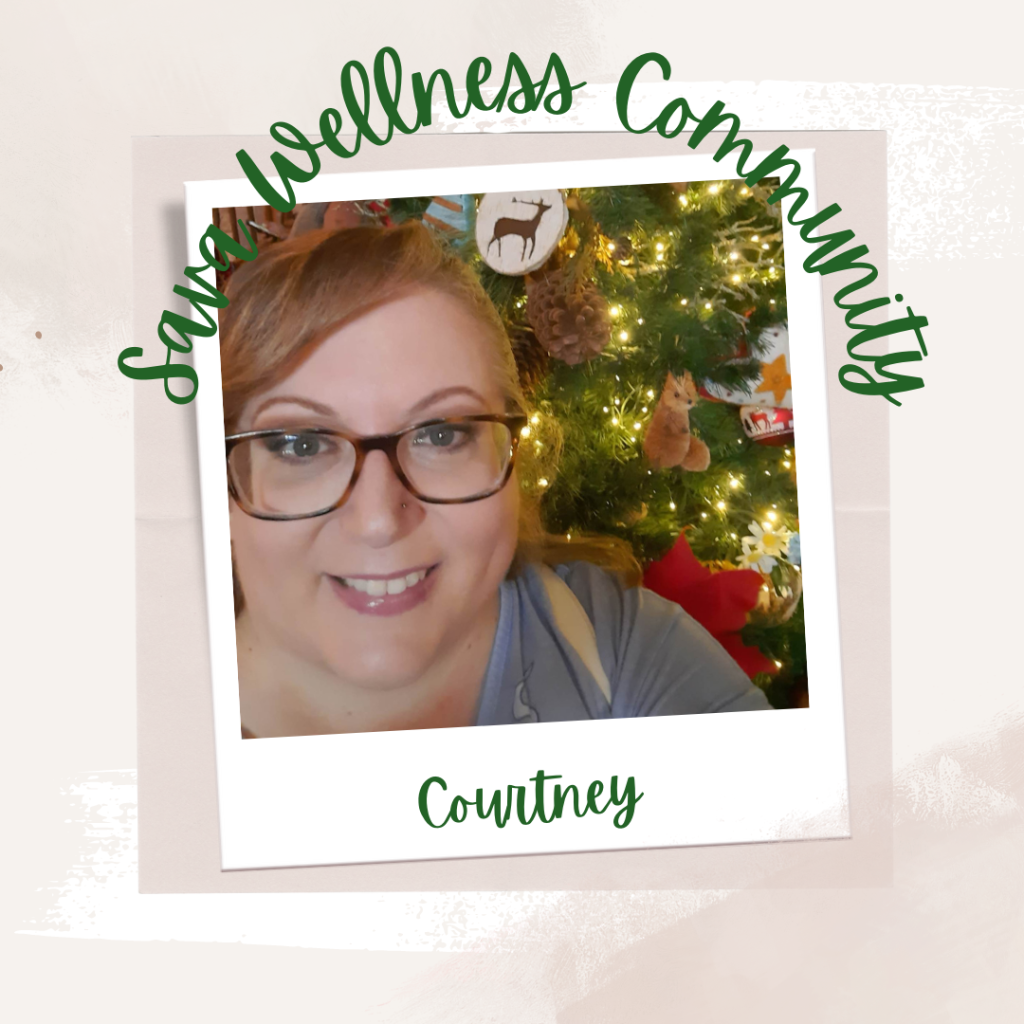 The words “Sava Wellness Community” are written in green cursive script over top of a polaroid picture. The polaroid picture is sitting on top of multiple layers of pink, cream, and white watercolour background. The polaroid picture has the name “Courtney” written in green under it. The picture is of Courtney smiling at the camera with a Christmas tree filled with woodland ornaments behind her. Courtney wears glasses and has her hair pulled back.